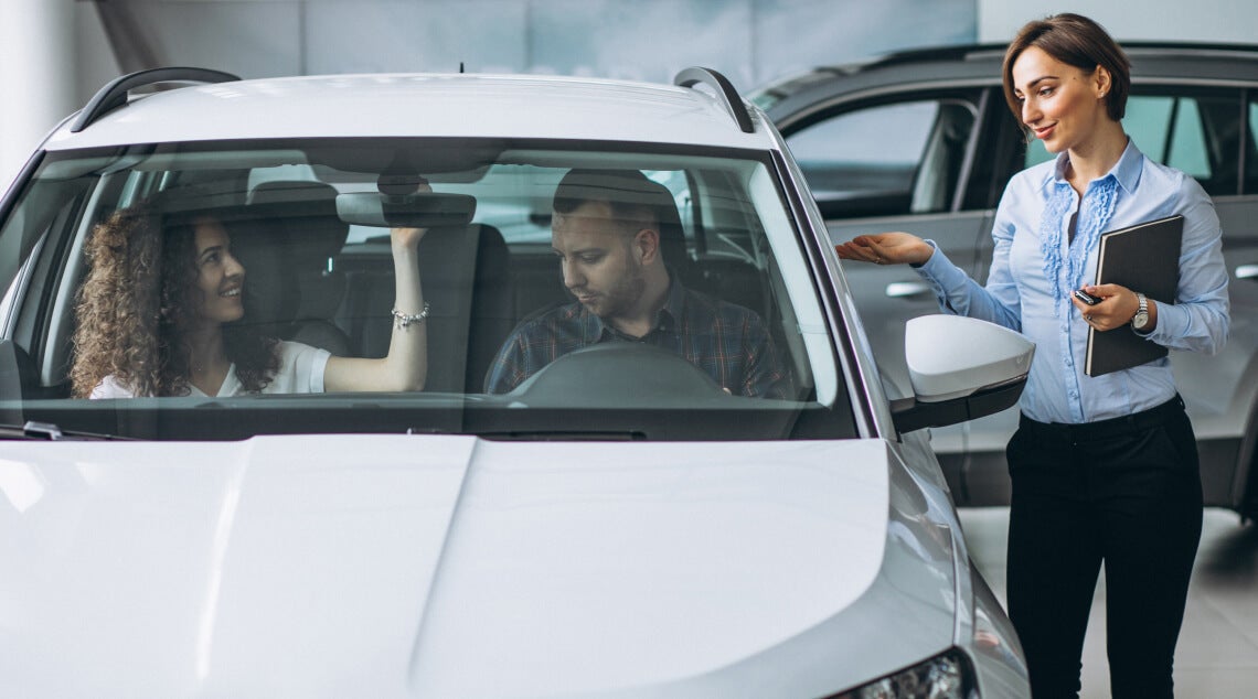 Dealership salesperson explains vehicle features to a couple seated inside a white SUV in the showroom.